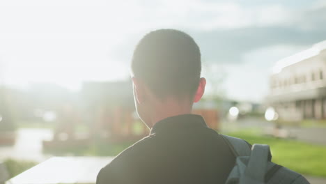 Back view of boy standing outdoors with backpack, sunlight casting soft glow around neck and shoulder, close detail of casual school bag with blurred greenery and benches in background