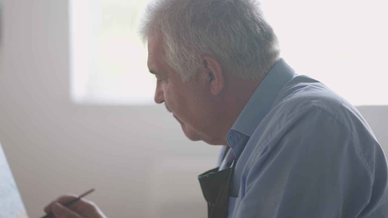 아프론 을 입은 노인 이 테이블 에 앉아서 브러시 로 그림 을 그린다