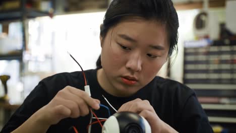 a young woman is intently working on connecting wires inside a device at a workshop. she demonstrates skill and concentration while engaging in hands-on electronics work.