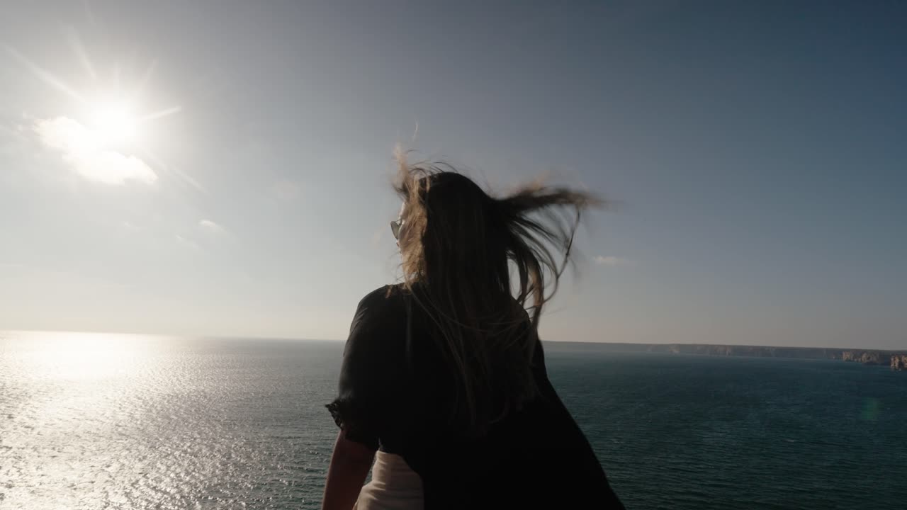 A woman stands on a rocky cliff in Algarve, Portugal, hair blowing in the wind as she gazes toward the glittering sea
