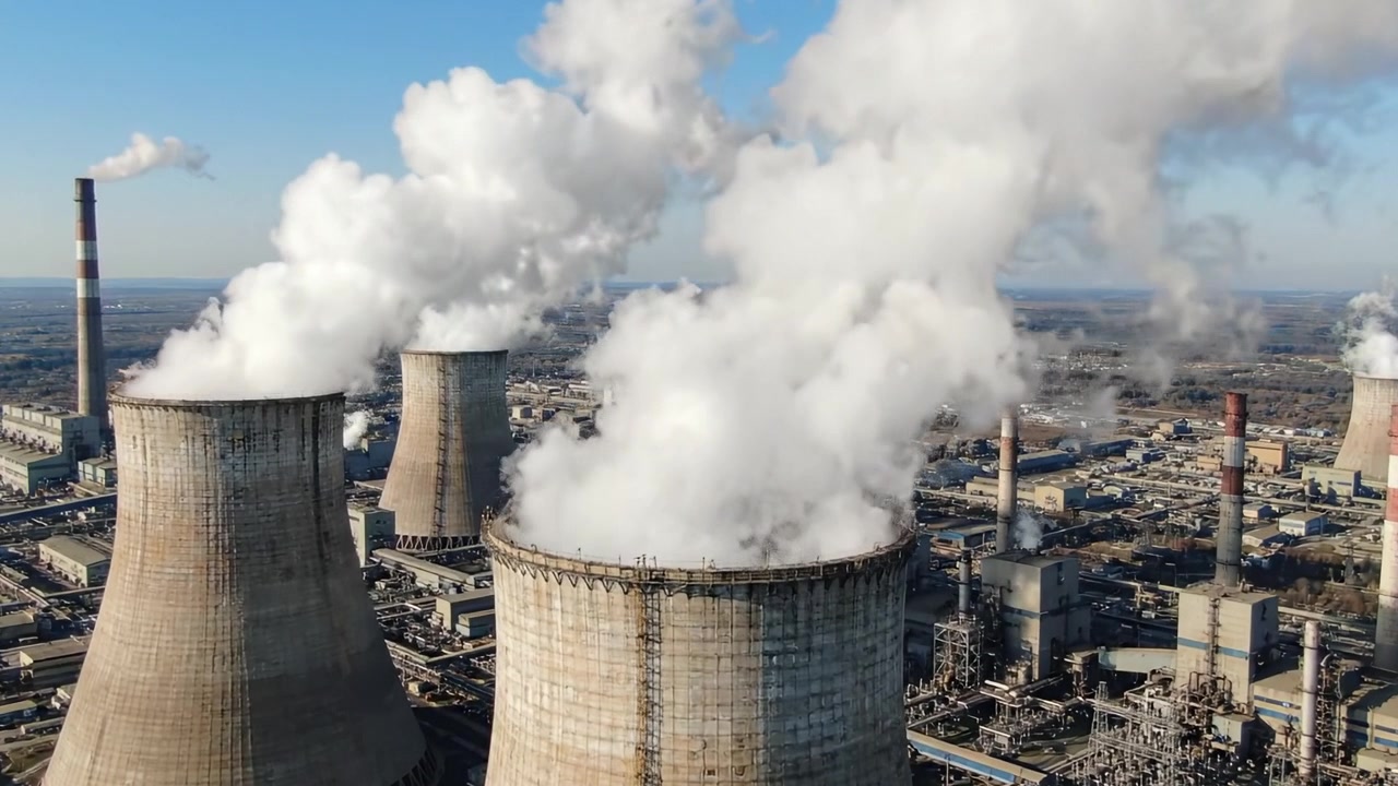 Centrale elettrica industriale che emette vapore: vista aerea delle torri di raffreddamento e delle strutture circostanti con fumo che sale contro un cielo azzurro limpido.