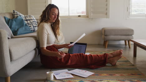 caucasian woman working from home, sitting on living room floor with laptop, coffee and paperwork