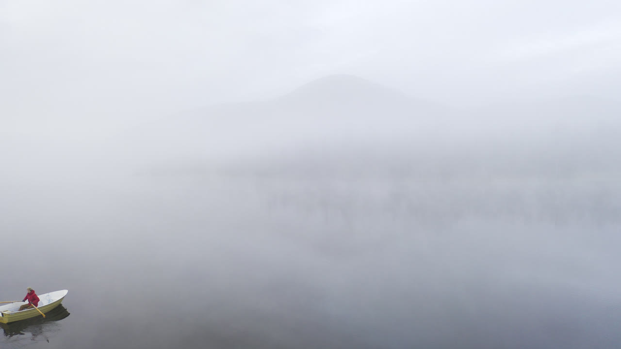 뒤쪽에 산이 있는 몽환적인 초현실적인 안개 풍경이 있는 호수에서 노 젓는 배를 탄 사람