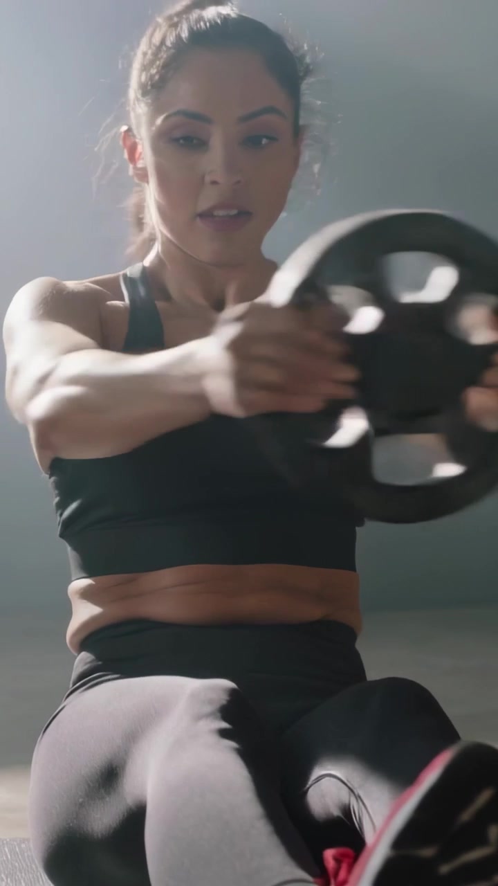Vídeo vertical: Início em ângulo baixo, mulher de roupa de ginástica girando no tapete segurando um peso para o core