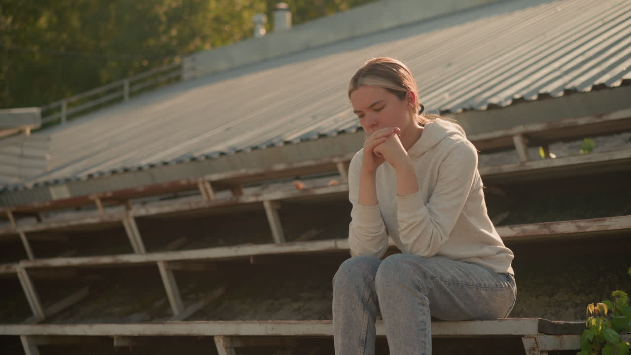 캐주얼한 후디와 청바지를 입은 여자가 농촌 스타디움의 관중석에 사려 깊게 앉아 있으며, 손이 함께 붙어 있고, 반사적인 표정으로