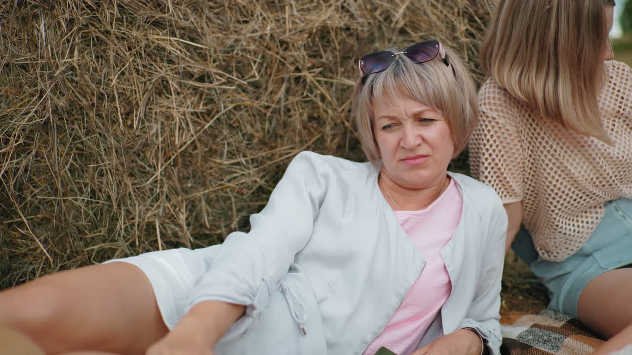 중년 여성은 개 줄을 들고 누워서 사려 깊게 보이지만, 그녀의 친구는 멀리서 쳐다보고, 열린 시골에서 풀 근처에서 편안한 순간을 보내고 있습니다.