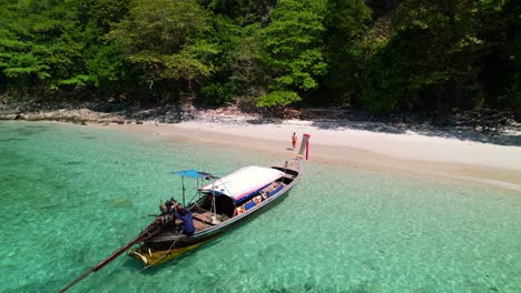 aerial drone circling a thai longtail boat anchored in the turquoise blue water of ko kai island in krabi thailand during a sunny day as a tourist stands on pristine white sand beach