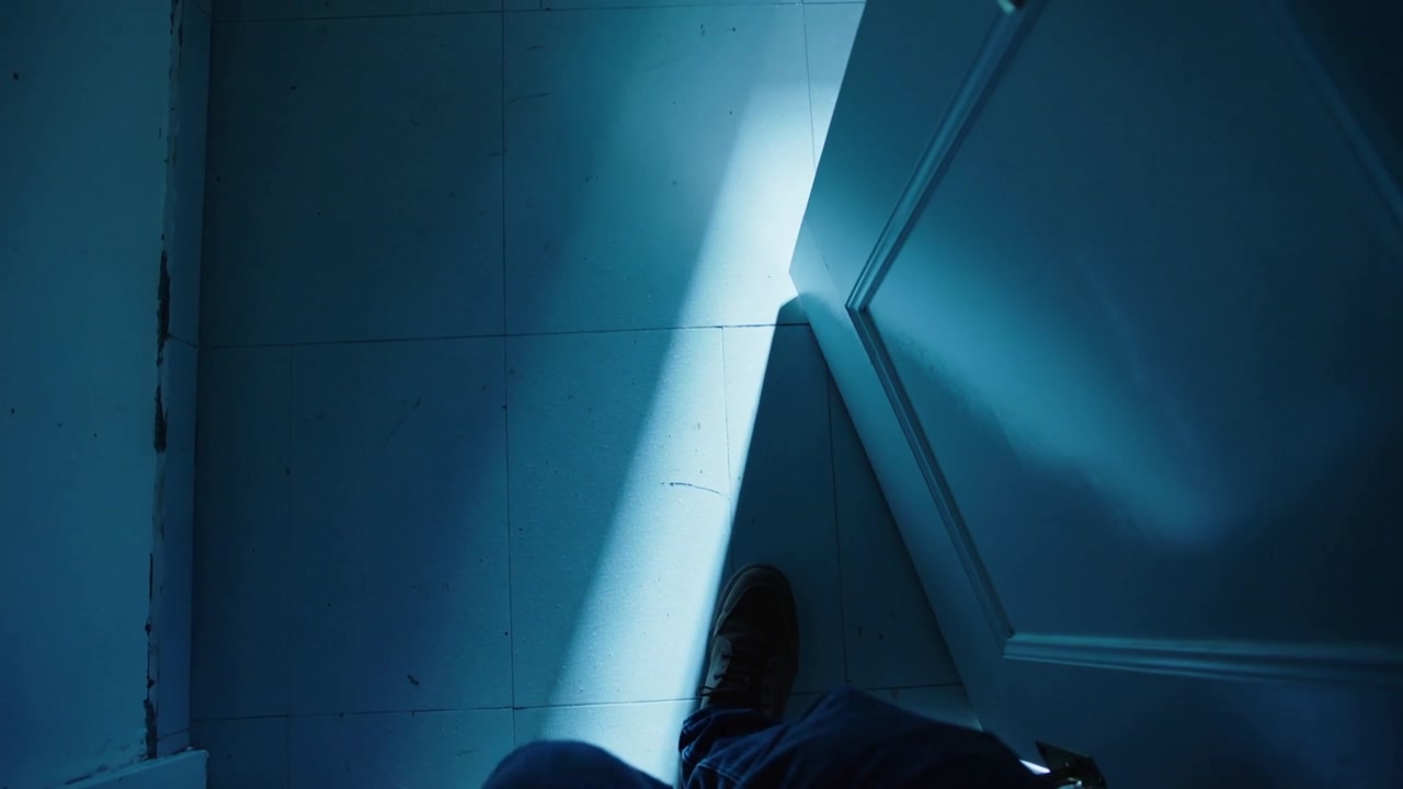 Vertical video: Stepping man in jeans brown shoes reacting to bluish beam under door on tiled floor