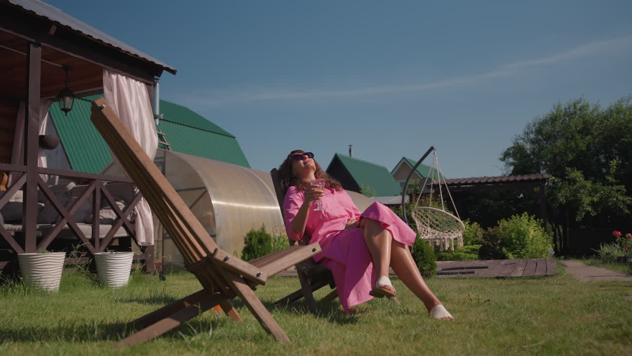 Woman In Pink Enjoying Sunny Garden, Fashionable Woman Relaxing By Cabin In Sunny Surroundings, Stylish Caucasian Woman Reclining Outdoors In Pink Dress With Sunglasses And Serene Atmosphere
