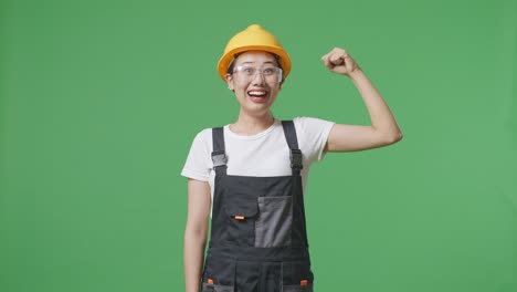 asian woman worker wearing goggles and safety helmet smiling and flexing her bicep while standing in the green screen background studio