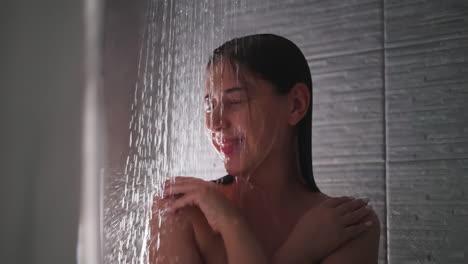 smiling woman stands in shower water closeup. happy relaxed lady spends time enjoying warm jets in home bathroom. body care procedures in washroom