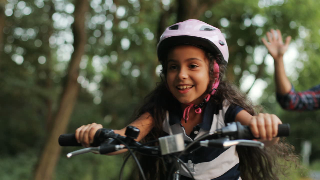 Closeup. Portrait of a little girl. Her dad is teaching her to ride a bike. He lets her go. Happy girl. Moving camera. Blurred background