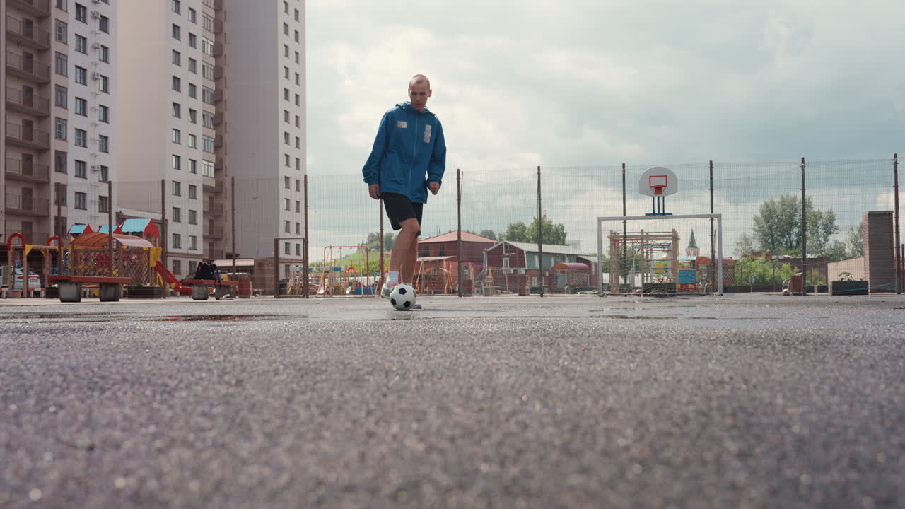 Solitary player training on wet asphalt, city court between apartment towers under overcast sky slow dribble and precise kick, closeup footwork near puddles, gritty atmosphere with worn ball