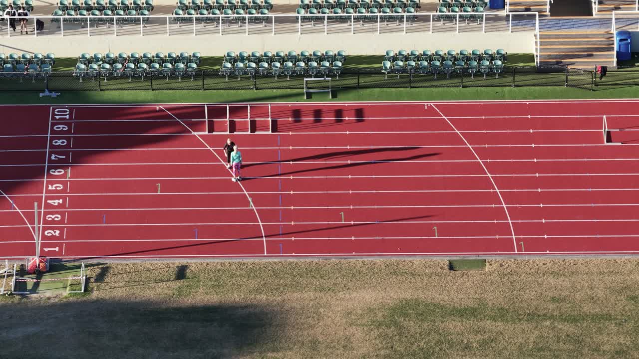 Two athletes practice on an empty athletics track under bright daylight, focusing on sprinting and hurdling techniques