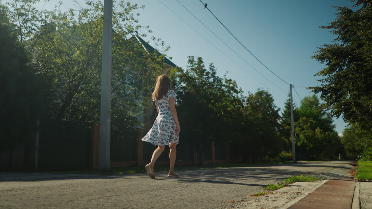 Back view of confident young adult in white floral gown walking along quiet suburban road while gently touching hair, surrounded by lush trees and low morning sun casting long shadows on pavement
