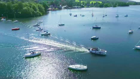 vista aérea giratoria elevada siguiendo un pequeño barco de crucero en el lago windermere en el puerto deportivo de bowness con yates en toda la escena en una mañana soleada de verano que muestra el problema de las algas en el lago