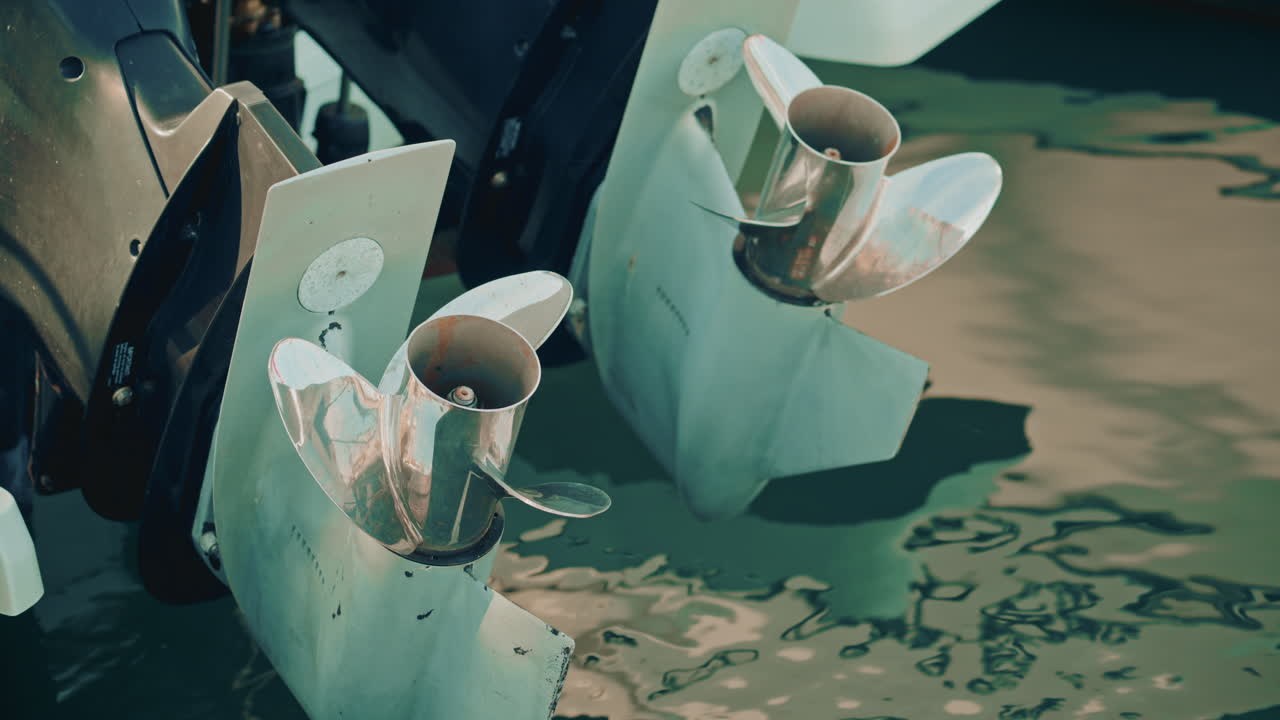 Two worn stainless steel boat propellers seen from above, rotating slightly in calm marina waters with abstract reflections