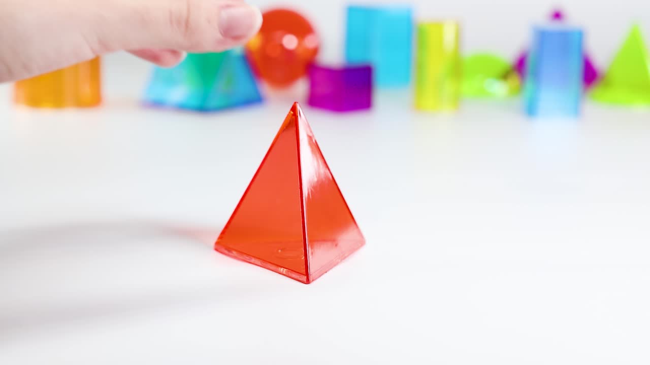 A hand arranges vibrant 3D geometric shapes on a white table, emphasizing educational play and spatial awareness