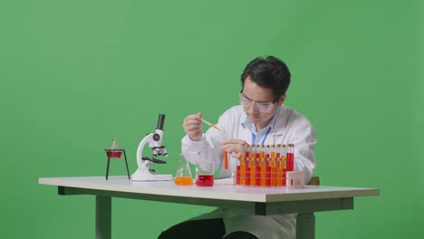 side view of asian man scientist making experiment with test tube and writing into notebook while working on the table with microscope in the green screen background laboratory