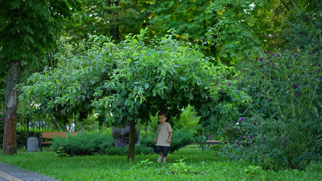 Charming boy runs by the green grass in the beautiful park. Kid runs to catch the small blue ball flying to him.