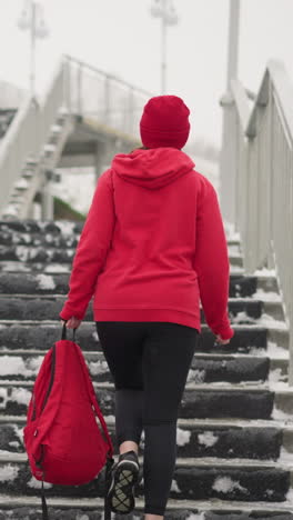 back view of woman in red hoodie walking up snowy staircase carrying red backpack, iron railings line stairs, snowy landscape surrounding, light poles adding wintry atmosphere to scene