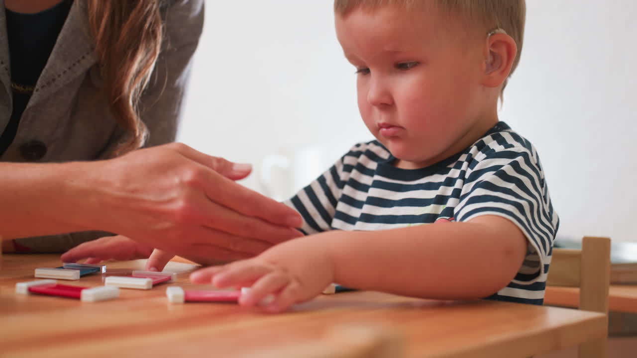 Un adulto lascia cadere dei blocchi su un tavolo di legno mentre un bambino con strabismo che indossa un apparecchio acustico li sistema con attenzione, maglietta a righe, espressione concentrata, guida di supporto che promuove un'istruzione inclusiva