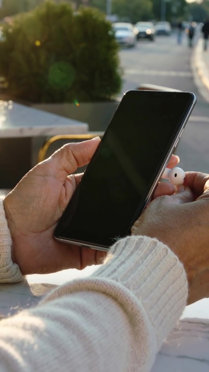 Video verticale: Il pollice che tocca, mani con polsini color crema che allineano un auricolare vicino a un telefono scuro per l'accoppiamento in un caffè