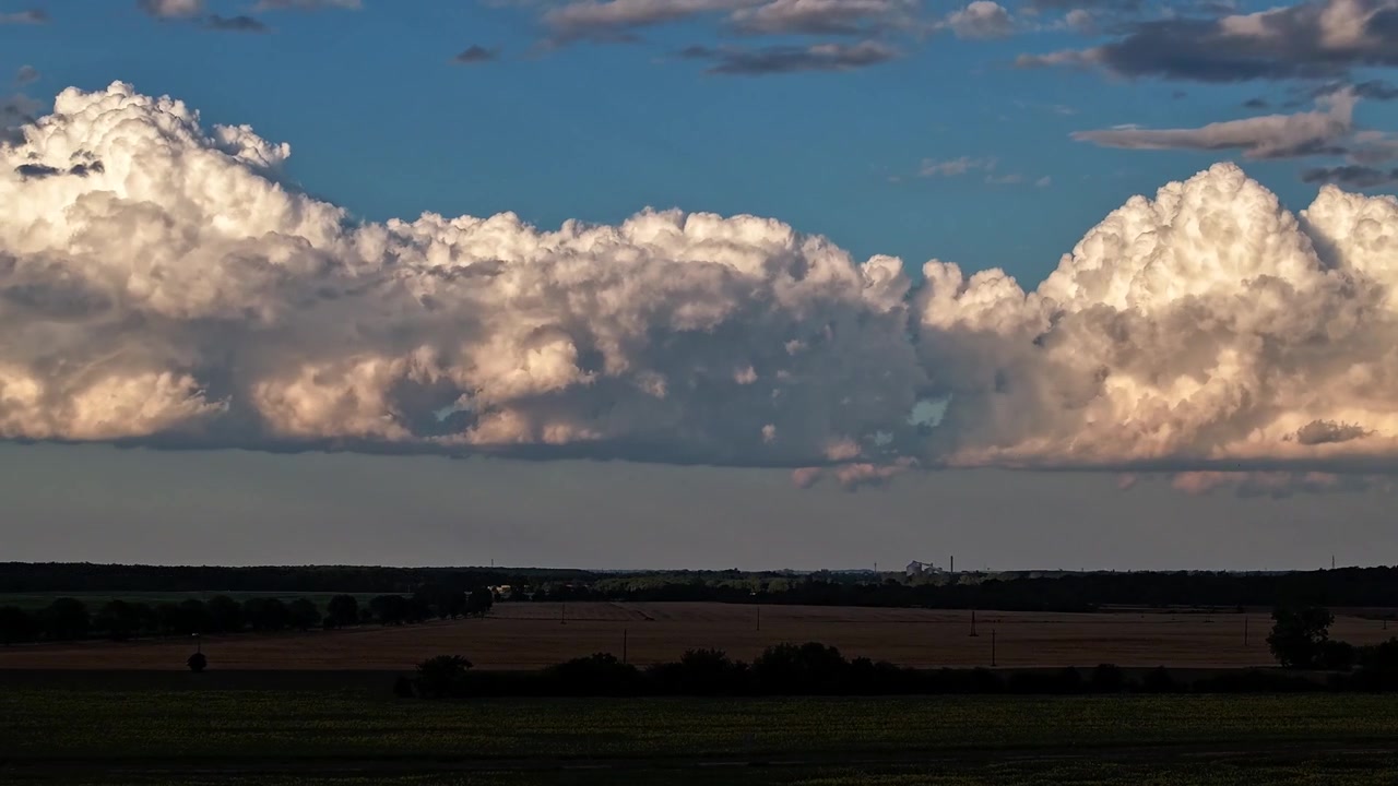 Timelapse of towering white cumulus clouds glow in warm evening sunlight above a peaceful rural landscape with open fields and a distant horizon.