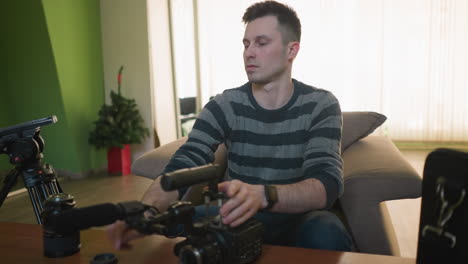 person sitting on sofa assembling camera with lens and microphone, focusing on filming process in home studio. preparing video equipment for content creation or filmmaking project