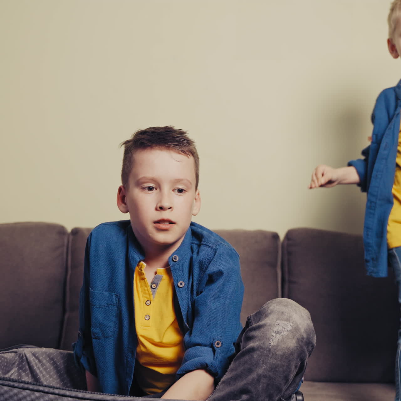 Two brothers are playing on the couch with travelling bag in the living-room. Elder boy is trying to lay himself into a grey suitcase at home.