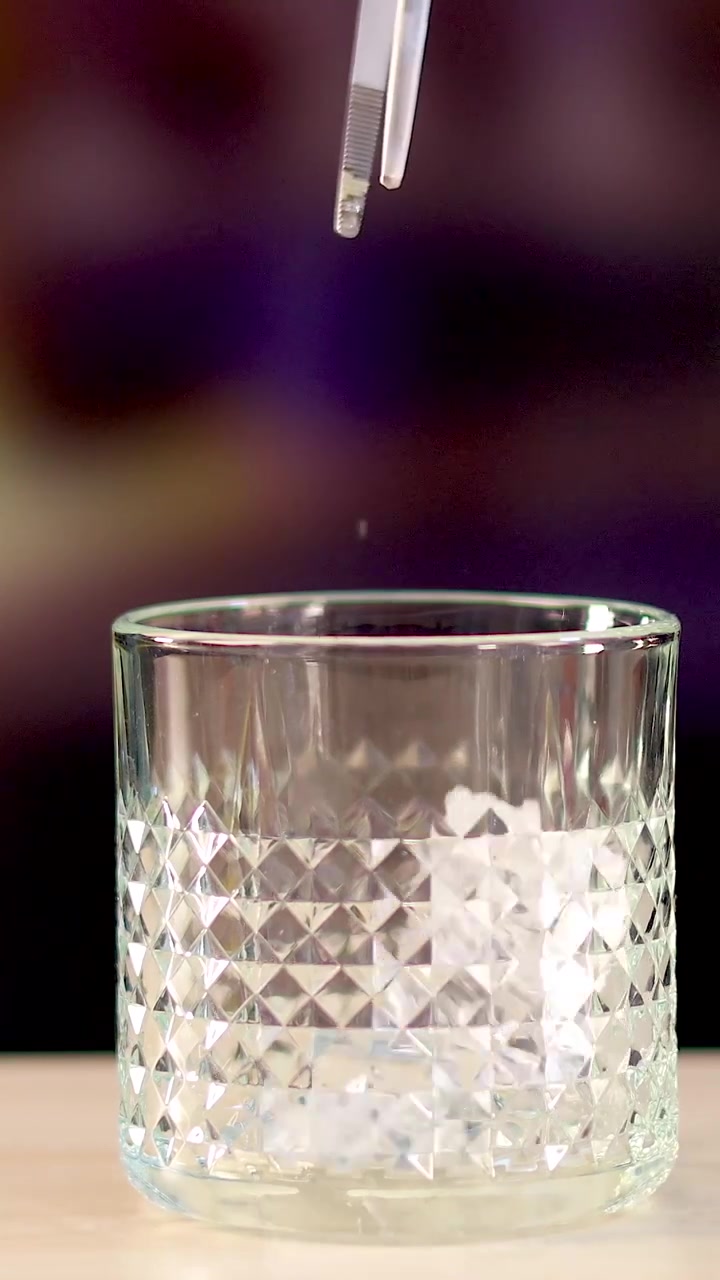 Cubetti di ghiaccio cadono in un bicchiere di cristallo sfaccettato su uno sfondo sfocato viola vibrante.