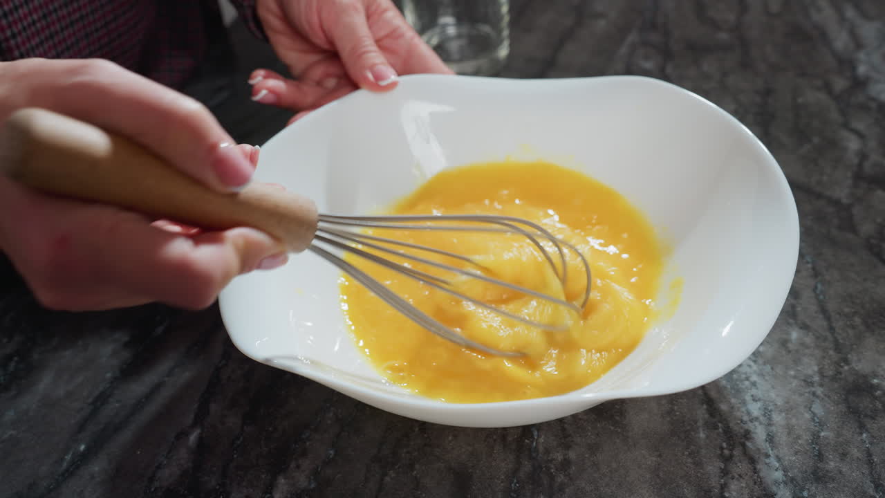 primer plano de un chef batiendo huevos en un cuenco blanco en una encimera oscura, la mano agita firmemente la mezcla, mezclando las yemas para hornear, el batidor de madera contrasta con el suave