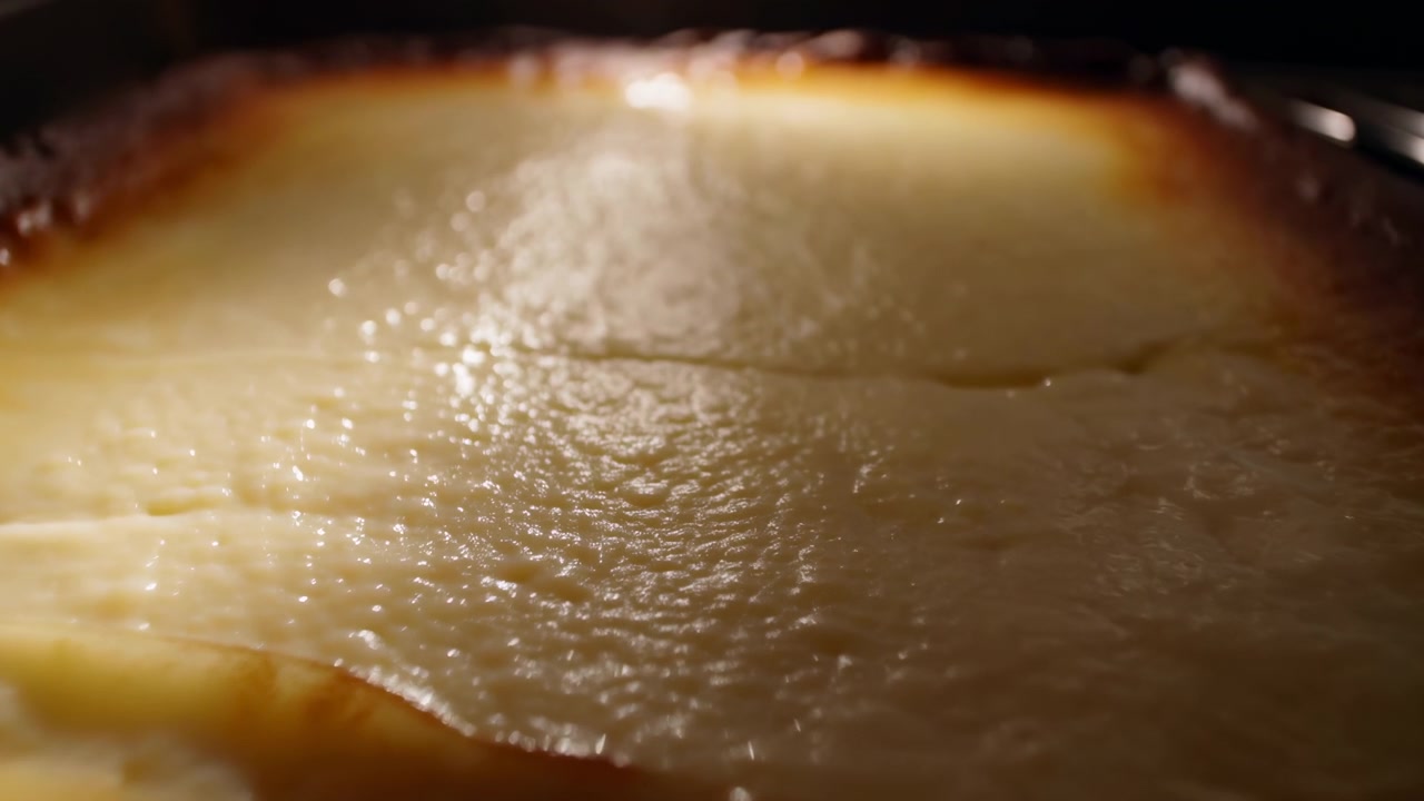 Vue rapprochée en contre-plongée de la surface d'un flan ou d'un cheesecake cuit dans un moule, montrant un centre jaune pâle brillant, une légère fissure centrale, de minuscules bulles, un bord caramélisé et une texture lisse à mesure que le dessert refroidit