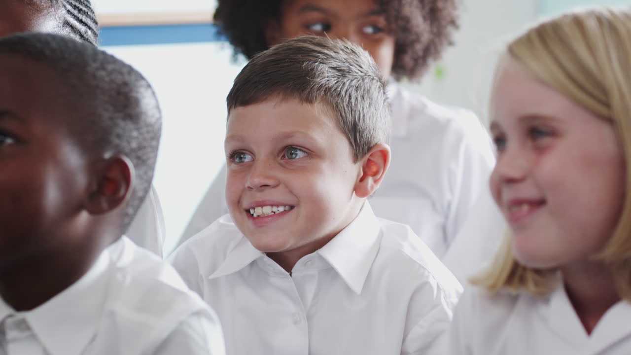 grupo de alumnos de primaria con uniforme sentados en el suelo escuchando al maestro en el aula