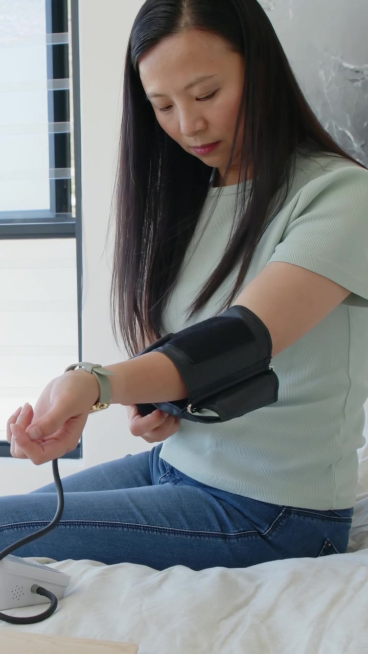 Video verticale: Donna asiatica applica un bracciale nero, preme il pulsante dell'unità sul letto, misurando la pressione sanguigna.