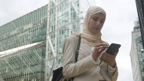 Muslim-Businesswoman-Checking-Messages-On-Mobile-Phone-Standing-Outside-Office-In-City