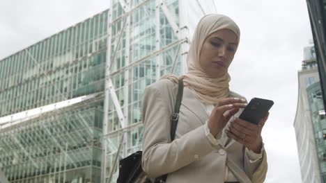 mujer de negocios musulmana chequeando mensajes en el teléfono móvil de pie fuera de la oficina en la ciudad