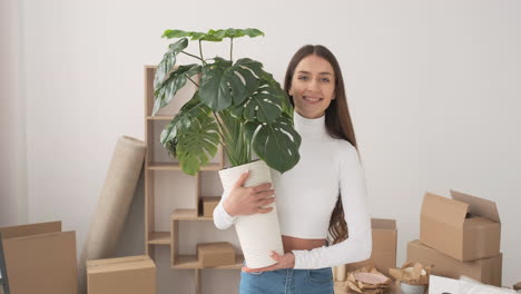 mujer feliz caminando con una planta en maceta y sonriendo a la cámara en un nuevo hogar