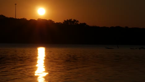 steady wide shot of a setting sun above the treetops with reflections in a lake