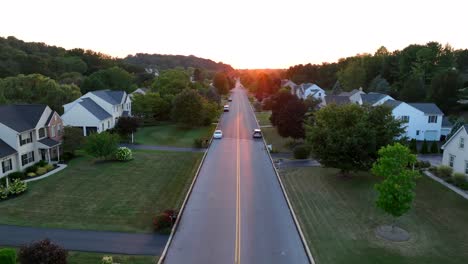 Aerial-view-of-a-suburban-street-at-sunset,-with-cars-on-the-road-and-houses-surrounded-by-trees