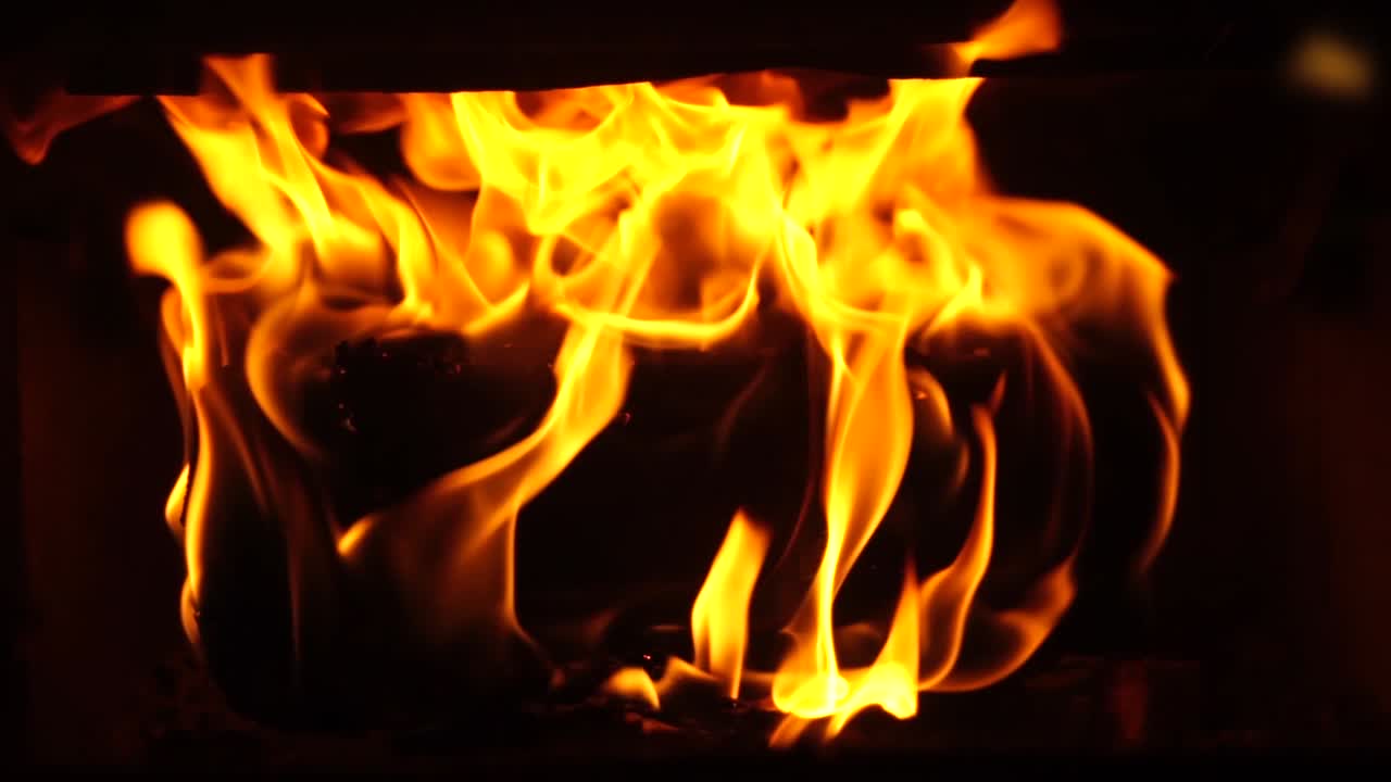 Gule flammer, der slikker og brænder på et enkelt brændestykke i en brændeovn af støbejern, orange og røde gløder, dyb sort kontrast i slowmotion