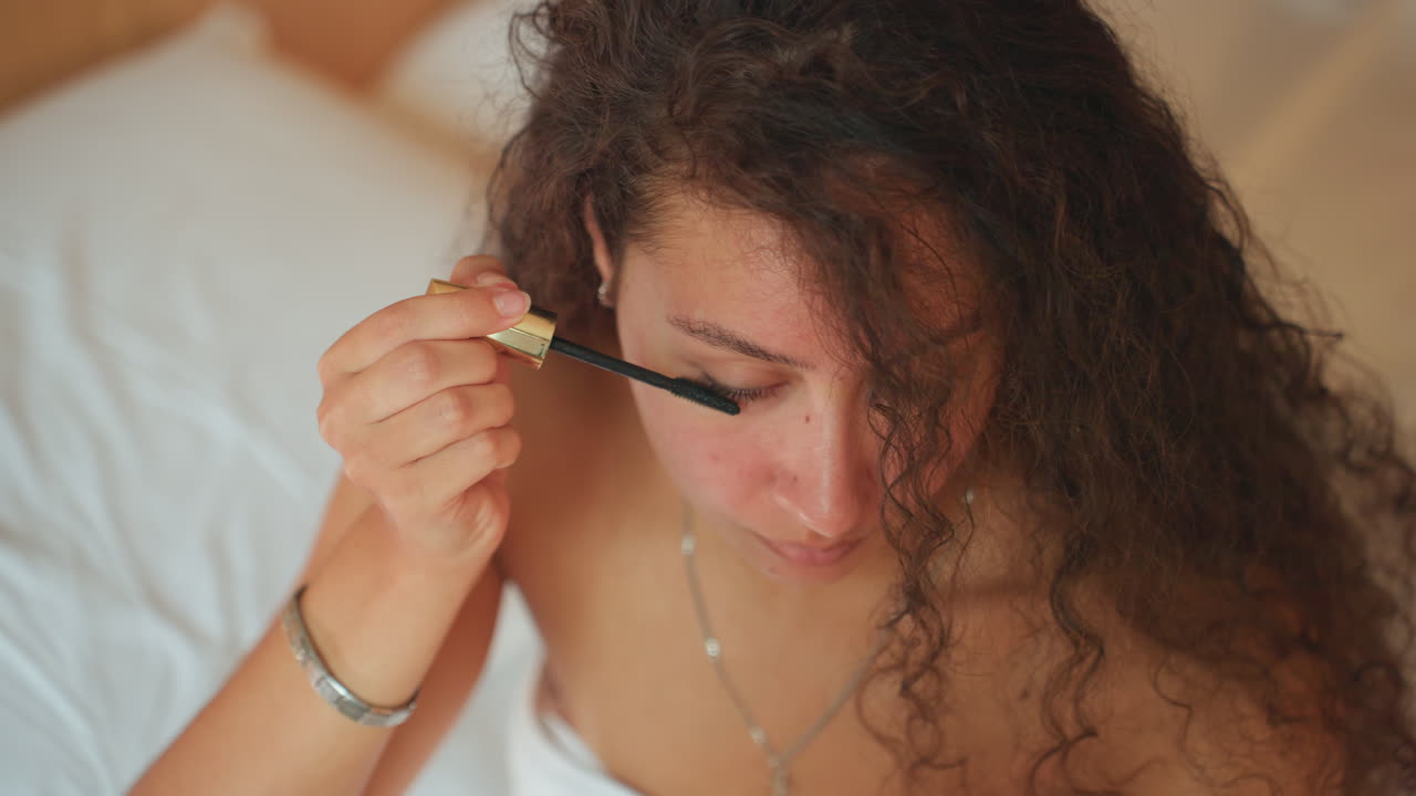 Woman applies mascara in hotel. Relaxed woman preparing for day ahead. Woman gently puts on makeup in cozy hotel room. Casually getting ready for day with makeup in comfortable hotel environment