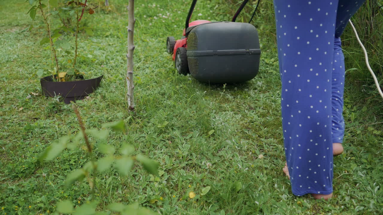 Legs of young woman mowing lawn by lawn mower near country house