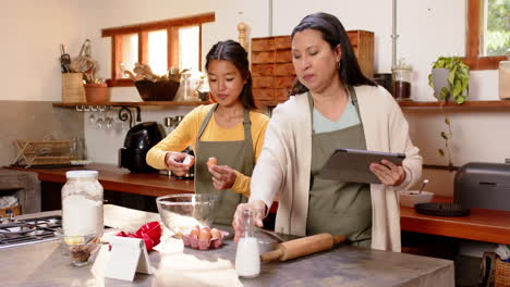 multiracial grandmother and young woman baking together in kitchen, using tablet for recipe, at home
