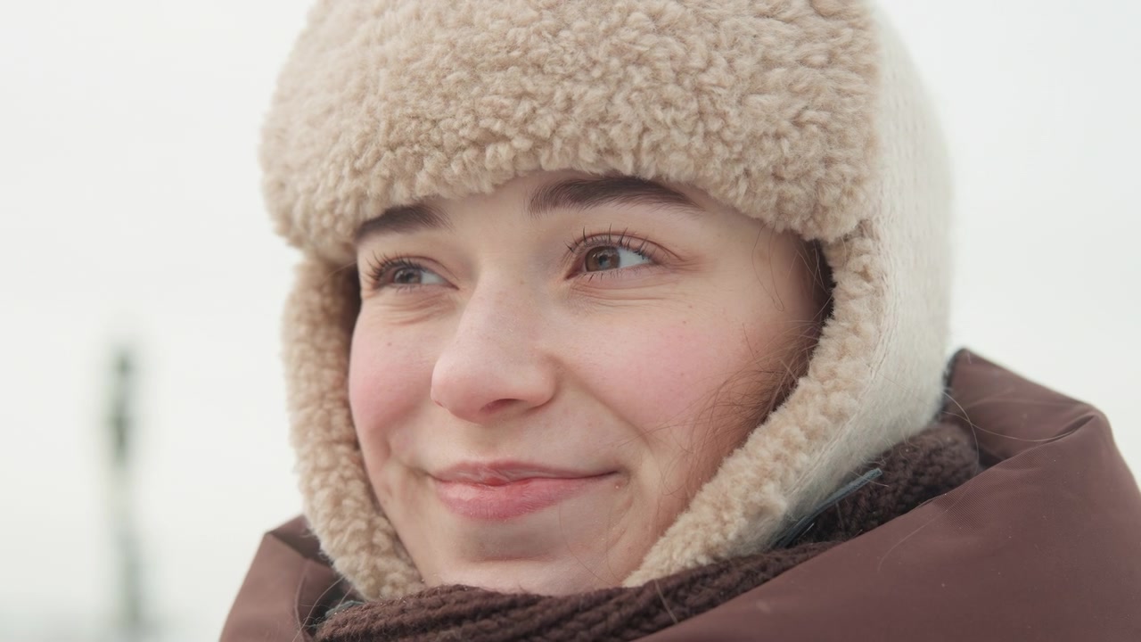 Ritratto all'aperto mostra una giovane donna con un cappello di pelliccia che solleva gli occhiali vicino alla bocca, poi si scioglie in un sorriso gentile con gli occhi che si chiudono e si aprono, catturando un'atmosfera invernale calma e uno stile casual.