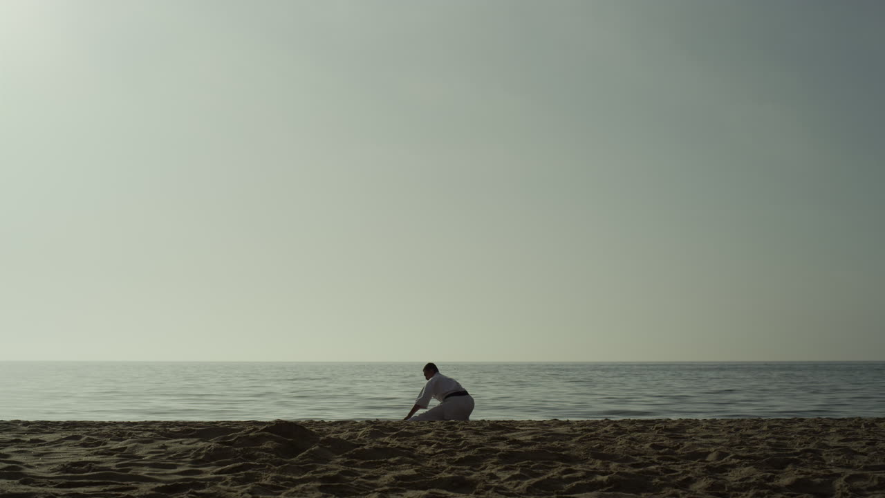 hombre deportivo haciendo calentamiento en cuclillas en la arena. luchador de karate estirándose en la playa.