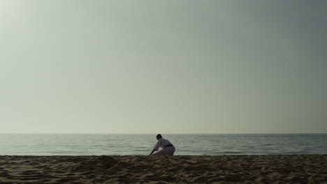 Hombre-Deportivo-Haciendo-Calentamiento-En-Cuclillas-Sobre-La-Arena.-Luchador-De-Karate-Que-Se-Extiende-En-La-Playa.