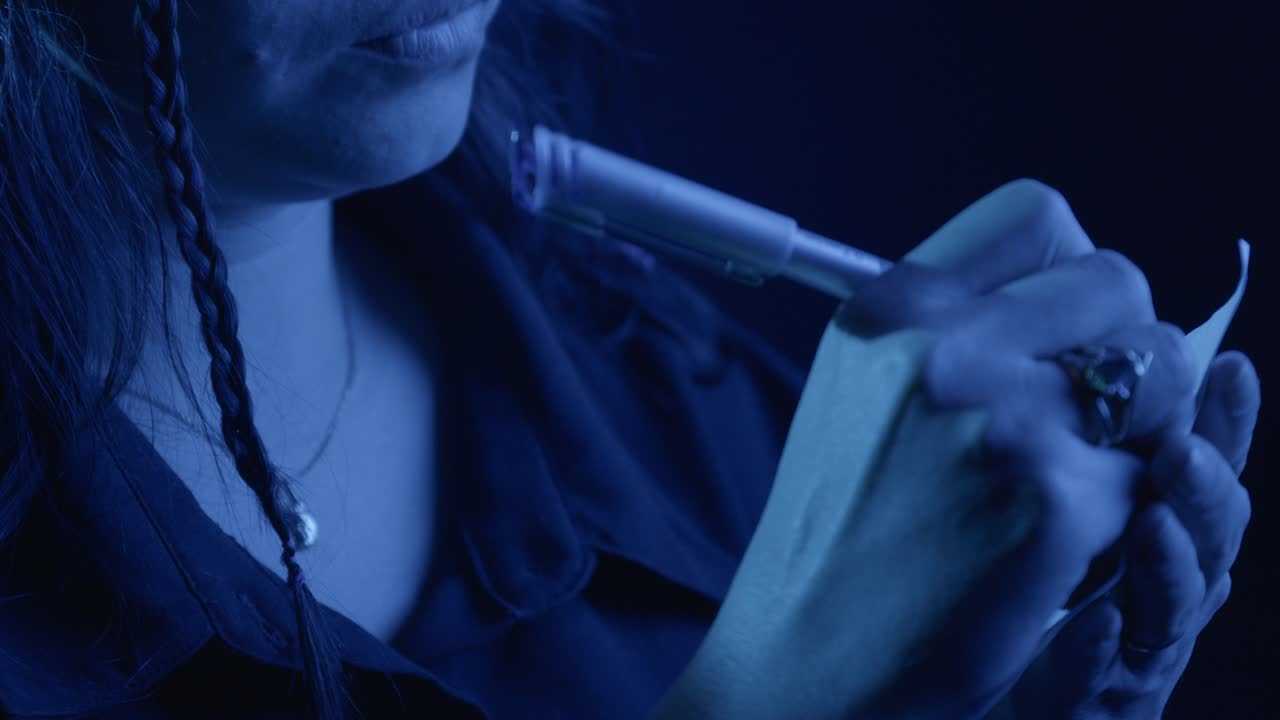 A woman pens notes in a moody blue close-up shot. The focus on her hand reveals intricate penmanship under low, dramatic lighting, creating an intimate and thoughtful atmosphere