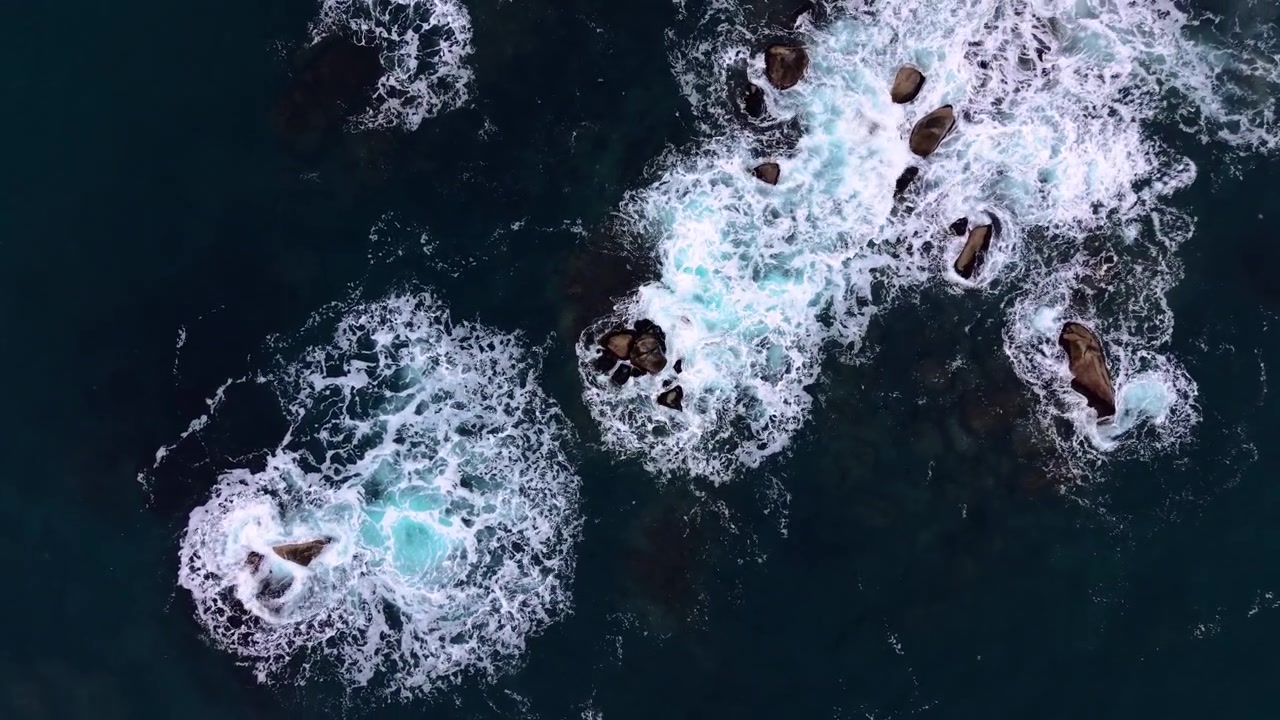 Aerial pattern of foamy waves breaking over rock. Overhead drone shot of dark blue sea, white foam and scattered boulder along the coast.