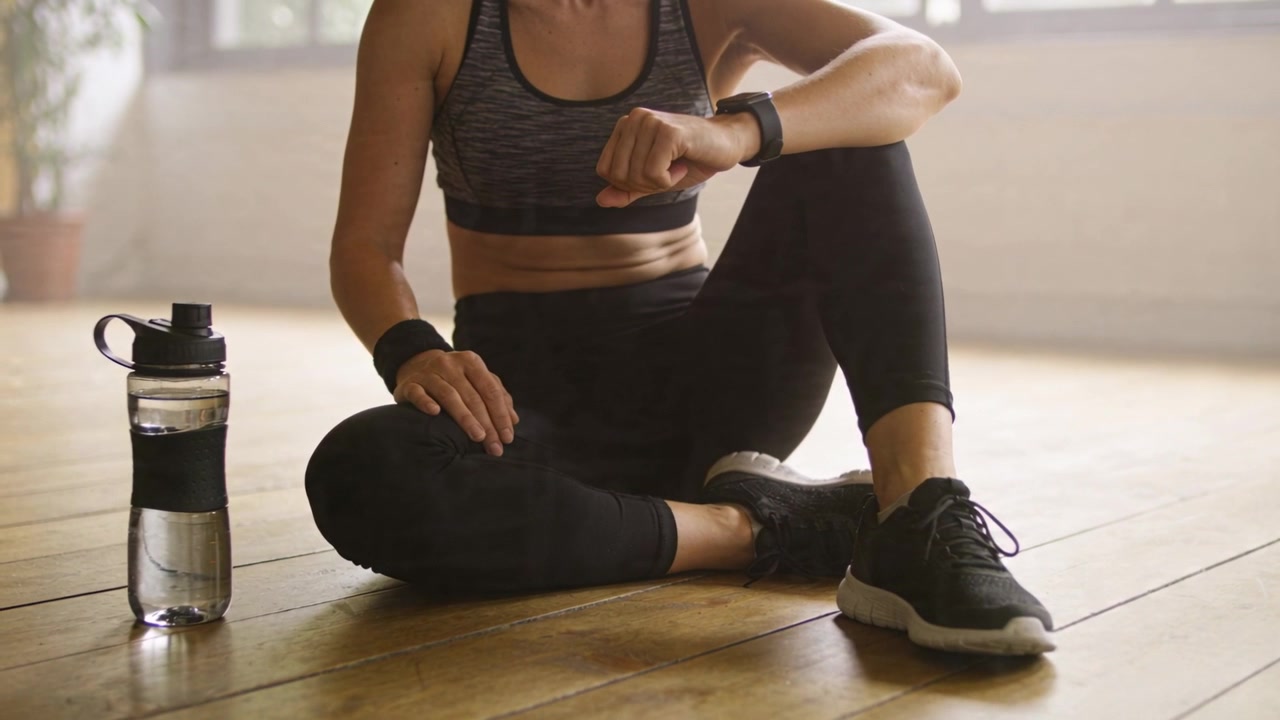 Sitting woman in sportswear, checking watch while resting on floor post-workout, water bottle left